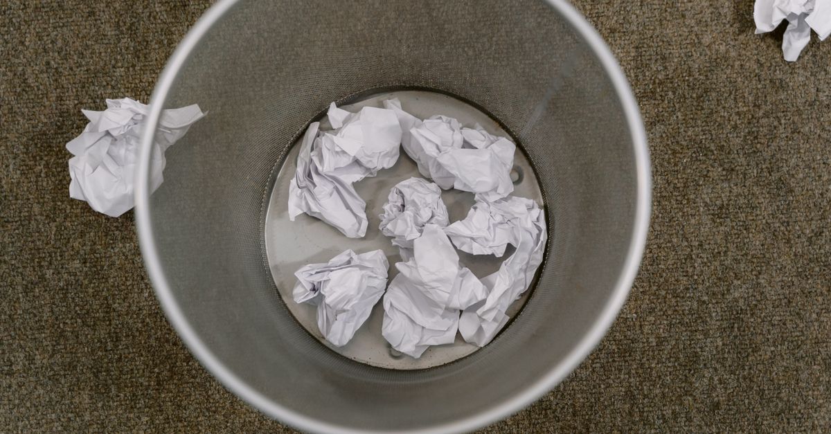 Overhead view of a trash can filled with crumpled paper on a carpeted floor.
